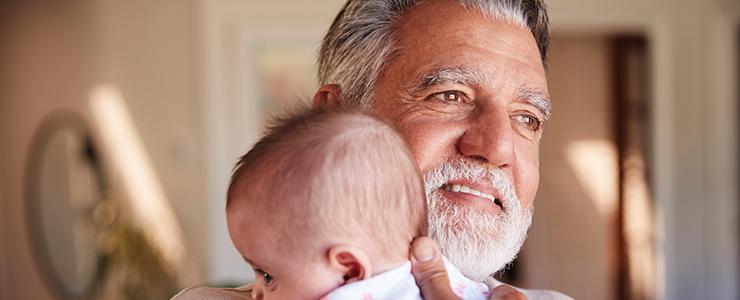 A smiling, white-bearded man gazes out on a sunny day, embracing an infant to his chest